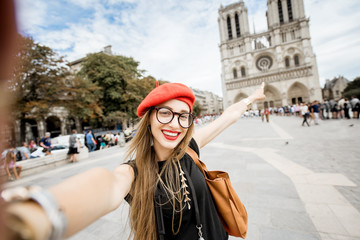 Young woman tourist in red cap making selfie photo standing in front of the famous Notre Dame...