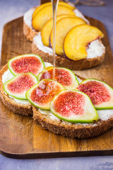Pouring honey on toasts with cream cheese topped with fresh ripe figs and peaches. Wholegrain rye bran bread on wood cutting board, grey concrete background.