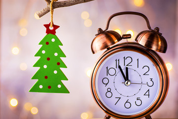 Vintage copper alarm clock showing five minutes to midnight, New Year countdown. Green christmas tree ornament hanging on branch. Glittering garland lights.