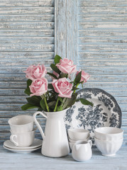 Pink roses in a white enameled pitcher, vintage crockery on blue wooden rustic background. Kitchen still life in vintage style. Flat lay