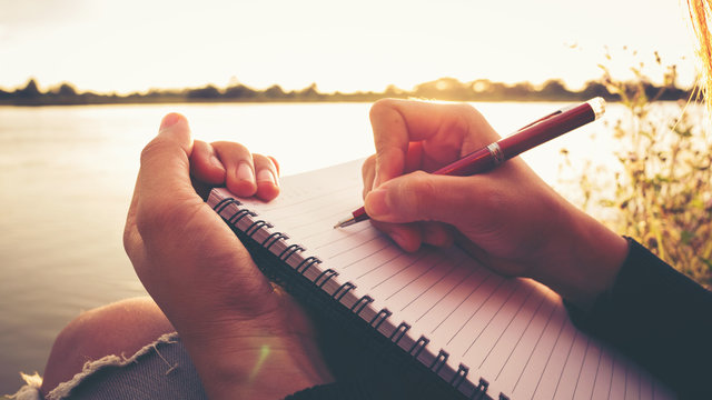 Close up hand of young woman with pen writing on notebook at riverside in the evening.