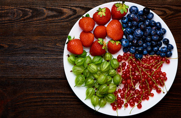 Garden berries on white plate. Strawberries, blueberries, currants, gooseberries on a wooden background. Top view.