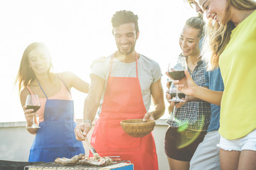 Happy students cooking and drinking wine at rooftop barbecue dinner - Best friends grilling meat...