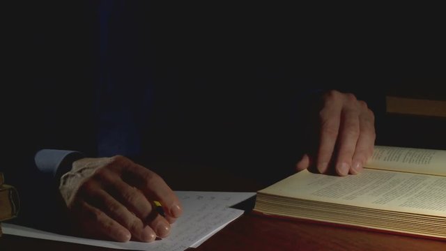 A person at night in the office at the table writes a pencil text on a sheet of paper, leafing through the book