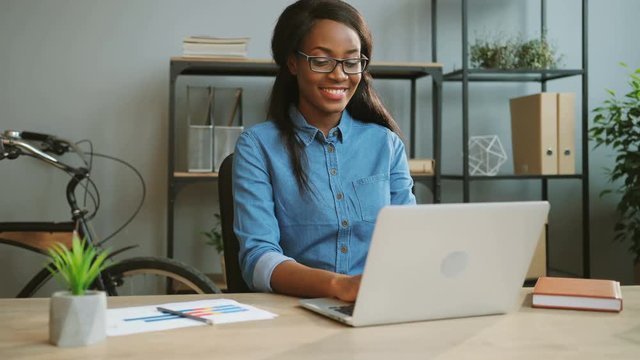 Young happy african american business woman in the glasses working on the laptop in the modern office while sitting at the table.