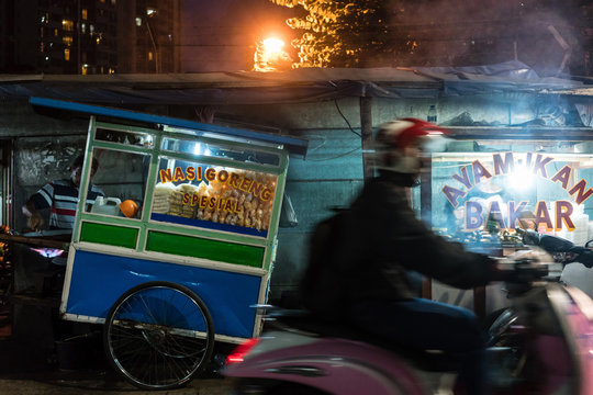 Side view of a man riding a scooter on a street with traditional food stalls in a poor district of Jakarta at night