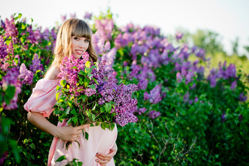The young beautiful pregnant woman is near a big  lilac bush with a lilac bouquet in her hand