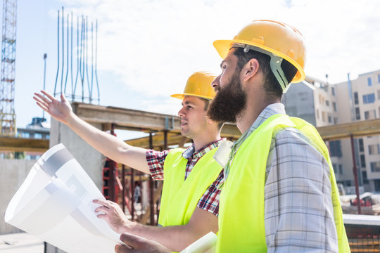 Side view of a skilled worker sharing with his colleague his innovative vision about a building while holding a blueprint outdoors on the construction site 