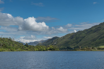 Fototapeta na wymiar Connemara, Ireland - August 4, 2017: Looking East over Pollacapall Lough from the grounds of Kylemore Abbeys shows blue water, a dam, blue sky with white clouds and forested green hills.