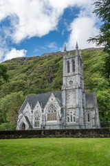 Connemara, Ireland - August 4, 2017: Small gray Neo-Gothic church on the grounds of Kylemore Abbey against rocky green hills and under blue sky with white clouds. Lateral view with dominant bell tower