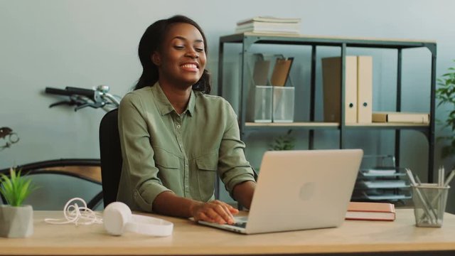 Young happy african woman working on the finance report using the laptop in the modern office, smiling and laughting during process.