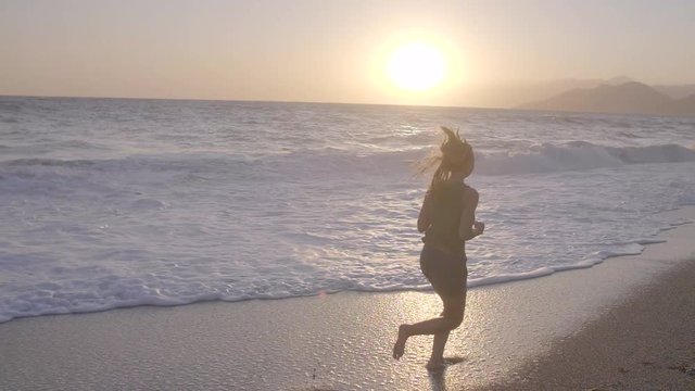 Young attractive woman with long blonde hair running or jogging along the sea shore at sunset. Slow motion shot of beautiful girl in shorts and tank. Happiness and freedom concept,