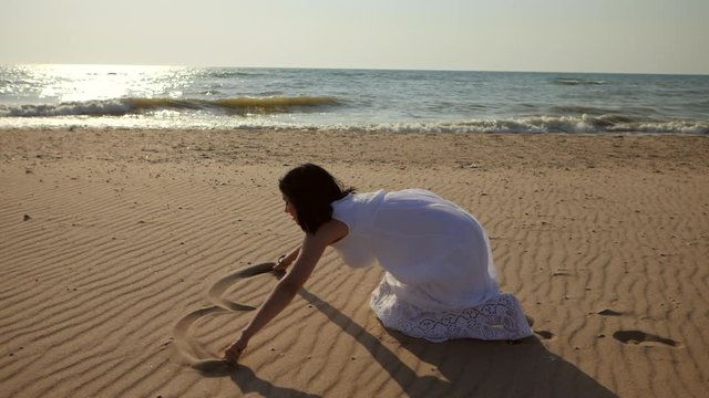 Beautiful caucasian Woman in long white dress sitting on beach and drawing hearts into wet sand on the shore. Love concept. Slow motion.
