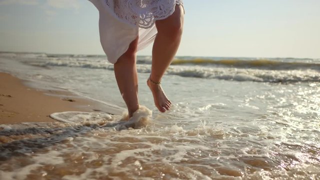 Legs of caucasian girl wearing white long dress and silver bracelets walking barefoot sand on sea beach, sun, lens, flare. Slow motion. Woman walks towards to camera