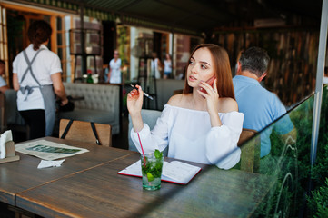 Portrait of a gorgeous young businesswoman talking on the phone and writing something down in her red notebook while sitting in a cafe.