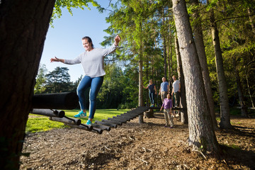 Friends Watching Woman Crossing Log Bridge In Forest