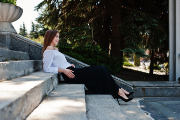 Good-looking young woman in white blouse, wide black pants and black classic high heels sitting on stairs and posing.