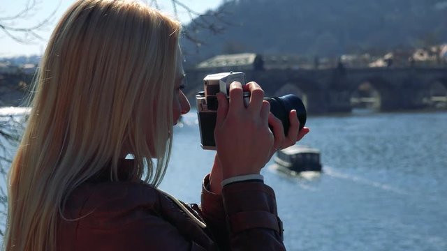A young attractive woman takes photos with a camera - face closeup from the side - a river and a bridge in the blurry background