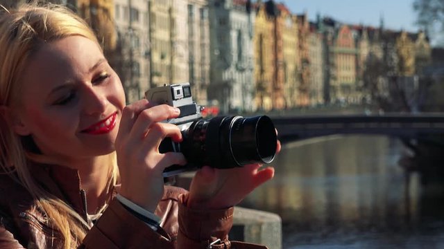 A young attractive woman takes photos, then smiles at the camera - face closeup - a river and a bridge in an urban area in the blurry background