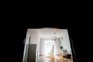 Look from the dark room at beautiful bride standing with her dress in the room