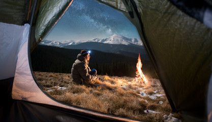 View from inside a tent on the male tourist have a rest in his camping in the mountains at night. Man with a headlamp sitting near campfire under beautiful night sky full of stars and milky way - obrazy, fototapety, plakaty