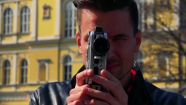 A young handsome man shoots a video with a camera - face closeup - a quaint yellow building in the blurry background