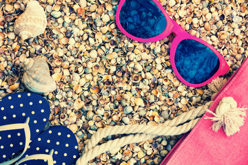 Beach scene. Woman accessories. Sunglasses, flip flop sandals and beach bag lying on sea coquina shells