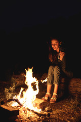 Young woman in geans and hoodie in the mountains. Young woman with fire on mountain at sunset. Girl standing near a fireplace, back view