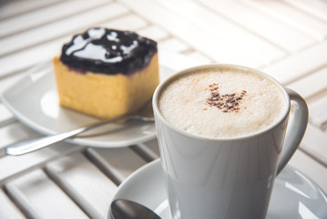 A cup of latte, cappuccino or espresso coffee on a white wood table and a Blueberry Cheesecake.