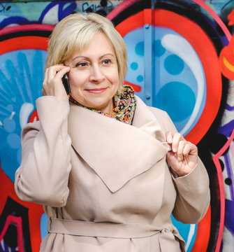 Beautiful Smiling Business Older Woman In A Light Beige Coat Talking On A Mobile Phone On The Graffiti Background