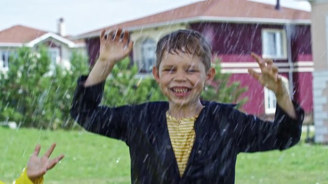 PAN with slow motion of happy boy with wet hair and his little brother in yellow raincoat laughing and jumping up and down on green grass during heavy rain