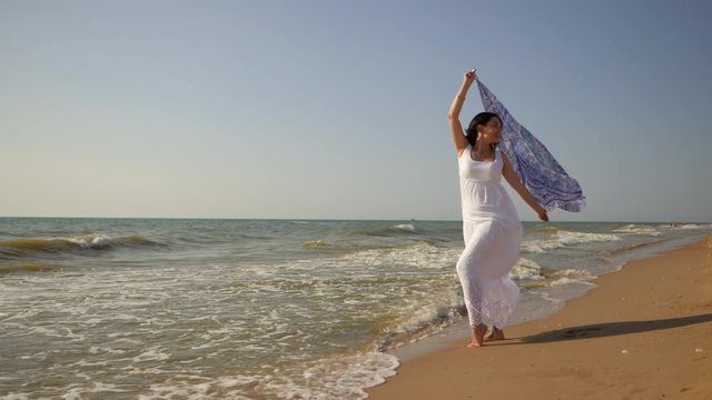 Gypsy young brunette girl wearing white maxi long dress standing near the sea or ocean windy beach with mandala silk scarf in hands. Bohemian clothing style. Boho lifestyle. Slow motion.