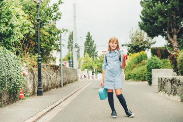 Outdoor portrait of funny little schoolgirl, wearing formal dress and backpack, holding lunch box...