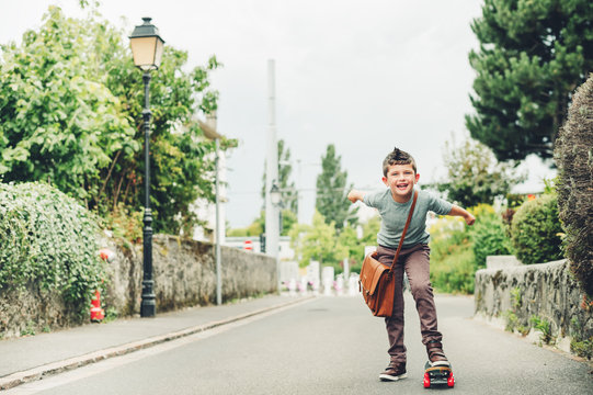 Outdoor portrait of funny little schoolboy wearing brown leather bag over shoulder, riding skateboard. Back to school concept. Film look toned image