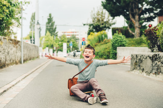 Outdoor portrait of funny little schoolboy wearing brown leather bag over shoulder, riding skateboard. Back to school concept. Film look toned image