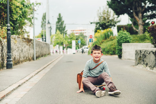 Outdoor portrait of funny little schoolboy wearing brown leather bag over shoulder, riding skateboard. Back to school concept. Film look toned image