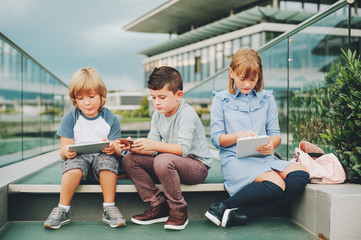 Group of 3 funny kids playing with tablet pc outdoors. Young children using electronic gadgets