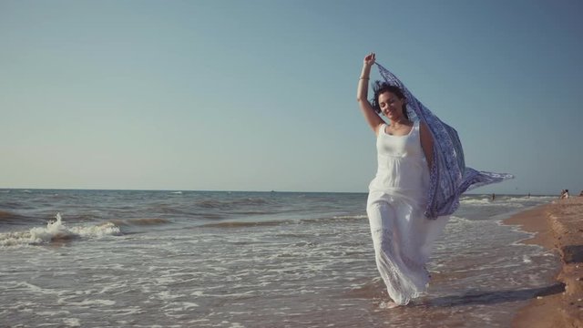 Gypsy young brunette girl wearing white maxi long dress running near sea or ocean windy beach with mandala silk scarf in hands.Bohemian style. Boho lifestyle.Slow motion.Film tonned