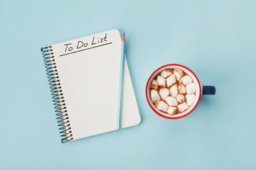 Cup of hot cocoa or chocolate with marshmallow and notebook with to do list on turquoise background top view. Christmas planning concept. Flat lay.
