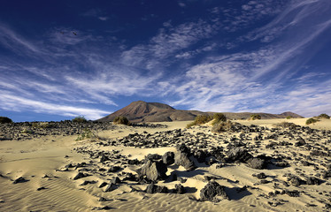 Wüstenlandschaft mit Sand, Steinen und Berge im Hintergrund unter blauem Himmel mit Windwolken.
