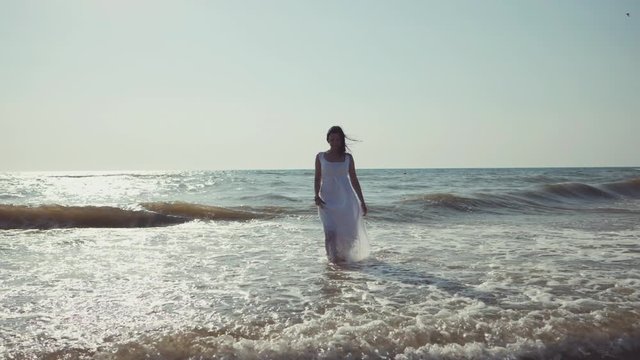 Gypsy young brunette girl in white maxi long dress walking on the waves in sea or ocean. Windy beach. Bohemian lifestyle. Slow motion. Film tonned