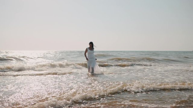 Gypsy young brunette girl in white maxi long dress walking on the waves in sea or ocean. Windy beach. Bohemian lifestyle. Slow motion. Film tonned