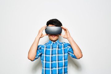 Waist-up portrait of young man wearing blue checked shirt using VR headset while standing against white background