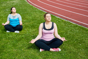 Calm females in activewear sitting in pose of lotus on green lawn