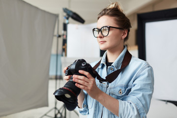 Waist-up portrait of talented young photographer in eyeglasses holding professional camera in hands and looking away pensively, interior of spacious photostudio on background