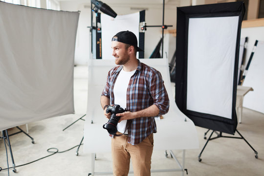Portrait of smiling bearded photographer wearing checked shirt and baseball cap looking away while taking short break from work, interior of spacious photostudio on background
