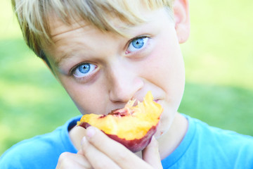 Portrait of child boy eating peach. Happy child in sun summer day. Kid with fruit in nature background.