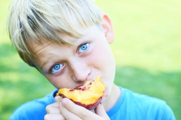 Portrait of child boy eating peach. Happy child in sun summer day. Kid with fruit in nature background.