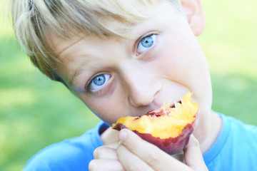 Portrait of child boy eating peach. Happy child in sun summer day. Kid with fruit in nature background.