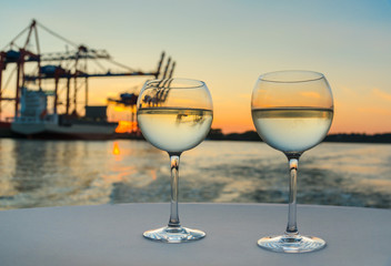 two glasses of cool white wine on white tablecloth with harbor structures and cargo ship in blurred background at sunset